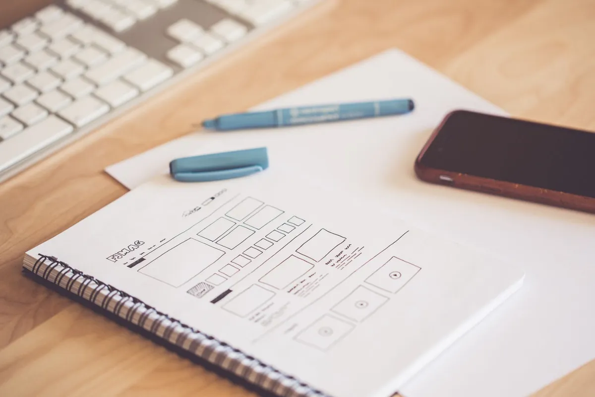 A notebook with hand-drawn wireframe sketches beside a smartphone and keyboard on a wooden desk, representing the early design stage before building screens in Figma