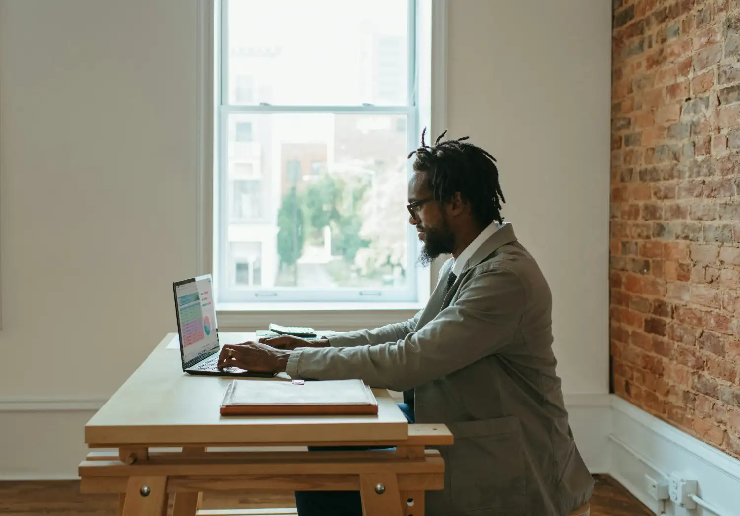 A professional freelance web designer working on a laptop at a wooden desk in a bright office studio with a brick wall, focusing on bespoke client solutions.